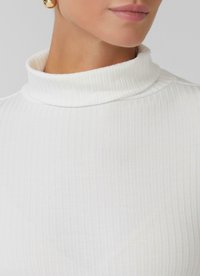 Gros plan sur une personne portant un pull à col roulé côtelé blanc et une grande boucle d'oreille en or, montrant ses lèvres et son cou.