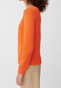 Donna che indossa un maglione lavorato a maglia arancione brillante con polsini e orlo a coste, abbinato a pantaloni beige, vista di profilo su sfondo neutro.