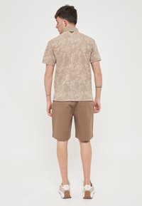 Maglietta polo color beige con motivo floreale e colletto, abbinata a pantaloni corti marroni e sneaker chiare, vista da dietro.