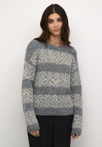 Pelēkā un krēmkrāsā rakstainā adītā džempera ar dimanta formām, apaļo kakla izgriezumu, garajām piedurknēm un mīkstu tekstūru, kombinācija ar tumšiem biksēm.