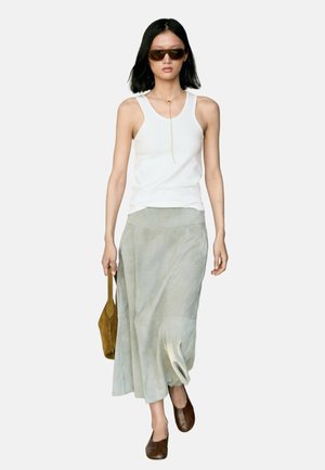 Donna con capelli neri che indossa occhiali da sole marroni, top bianco senza maniche, gonna grigio chiaro, scarpe marroni e tiene una borsa marrone.