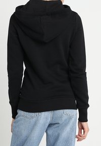 Persona vestida con una sudadera negra sencilla y jeans azul claro, vista desde la espalda contra un fondo blanco liso.