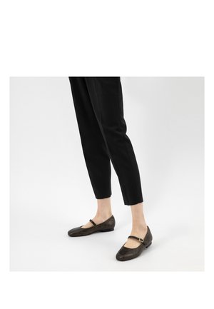 Jambes portant un pantalon noir raccourci et des ballerines Mary Jane marron foncé avec une bride unique et un talon bas sur fond blanc.