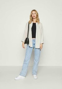 Camisa oversized beige com riscas finas e um bolso, usada sobre uma blusa preta, combinada com jeans azul claro e ténis brancos. Bolsa preta.