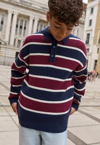 Băiat îmbrăcat cu un pulover tricotat în dungi, de culoare navy, burgund și alb, cu guler, stând afară lângă clădiri de stil clasic.