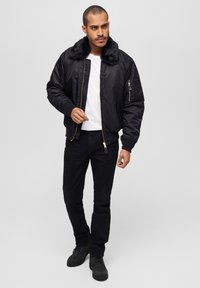 Chaqueta bomber negra con cuello de pelaje sintético, cierre en la parte delantera y bolsillos laterales, combinada con una camiseta blanca y jeans negros.