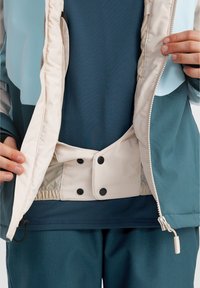 Veste bleu canard et crème avec une doublure en maille, fermeture à pression et taille élastique. En dessous se trouve un haut bleu foncé et un pantalon texturé assorti.