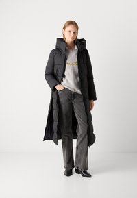 Lang svart dunjakke med hette og kviltet design, iført over en grå genser og grå jeans. Sorte ankelstøvler fullfører antrekket.