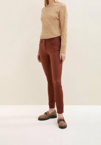 Světle hnědý pletený svetr s žebrovanými manžetami, kombinovaný s úzkými džíny v barvě rezavého oranžového. Hnědé mokasíny s černými akcenty dotváří celý outfit.