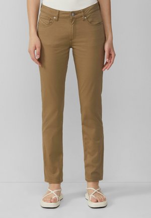 Pantalons en tissu extensible marron, avec une coupe slim, des poches avant et une fermeture à bouton, associés à des sandales blanches à lanières.