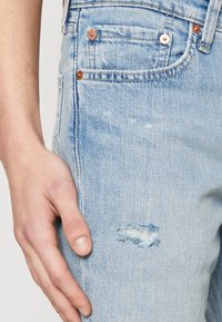 Světle modré džíny z denimu s vyřezanou nášivkou na stehně. Obsahují měděné kování a detailní záběr ruky odpočívající na látce.