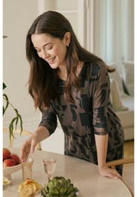 Brunt mønstret tøj med trekvartlange ærmer. En kvinde står ved et bord med æbler, en artiskok og gennemsigtige glas.