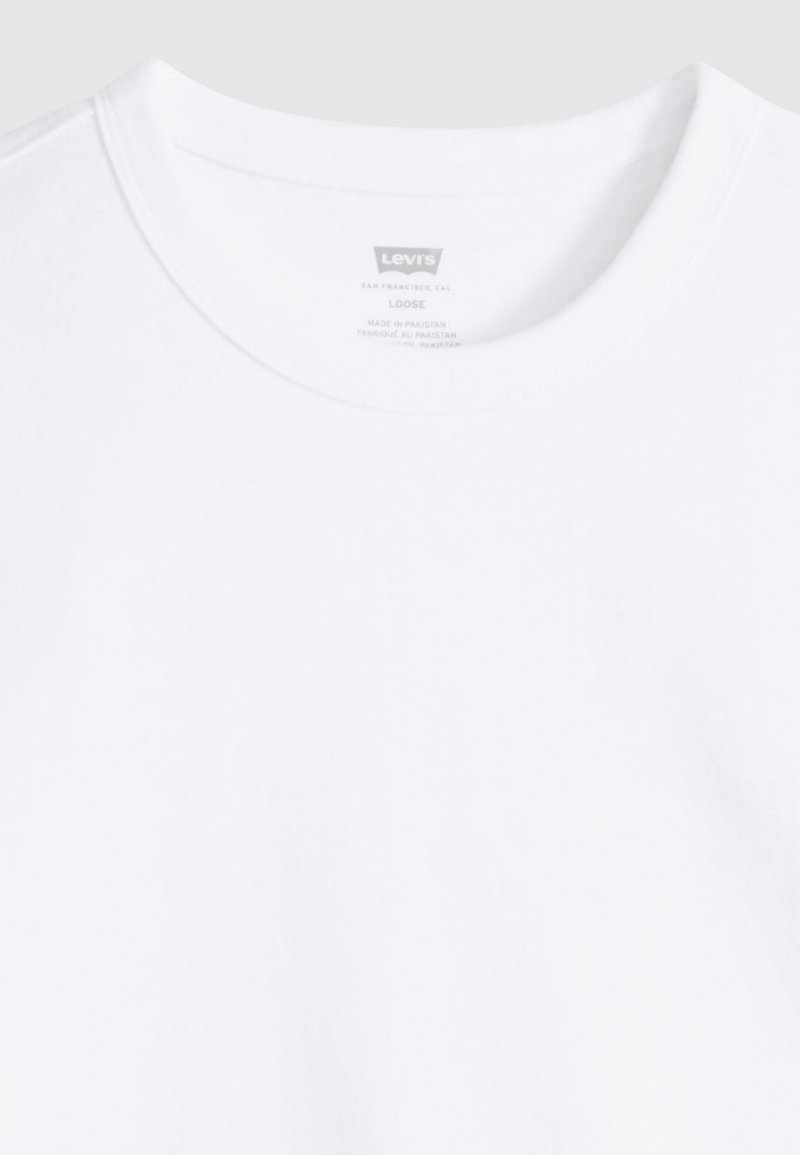 T-shirt blanc à col rond en coton, avec une coupe ample. Étiquette visible au niveau du col indiquant le nom de la marque et les instructions d'entretien.