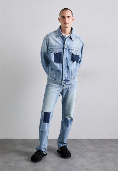 Denim jacket with light blue color, featuring dark blue patchwork accents. Coordinating light blue jeans with dark denim knee patches. Black shoes.