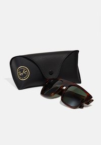 Ochelari de soare Ray-Ban cu ramă de broască țestoasă și lentile verzi, alături de un toc din piele texturată neagră, cu un logo auriu pe față.