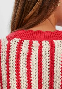 Czerwono-kremowy sweter w paski z rypsowym kołnierzykiem i falbaniastym wykończeniem. Ma różowy guzik z tyłu. Widoczna tekstura dzianiny.