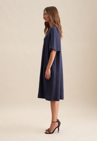 Navy-blaues Leinenkleid mit lockerer Passform, kurzen Ärmeln und knielangem Saum. Kombiniert mit schwarzen, hochhackigen Sandalen, die minimales Hardware-Design aufweisen.