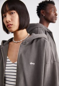 Grå zip-up hættejakke med en blød tekstur og et broderet "obey" logo på brystet, parret med en stribet tanktop nedenunder.