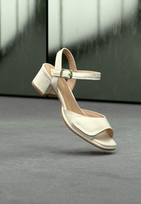 Άσπρο σανδάλι με ανοιχτά δάχτυλα, τακούνι τύπου block και λουράκι στον αστράγαλο, τοποθετημένο σε γκρι επιφάνεια με θολό σκοτεινό φόντο.