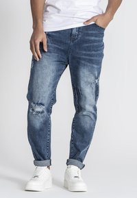 Jeans de mezclilla azul claro con desgastes, un corte relajado y puños enrollados, combinados con zapatillas blancas sobre un fondo liso.