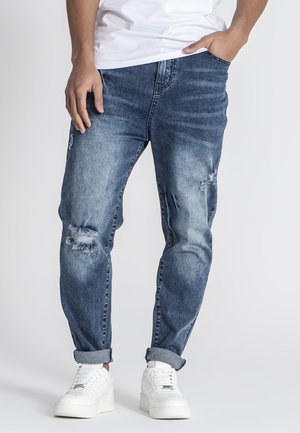 Lichtblauwe denim jeans met beschadigde scheuren, een relaxte pasvorm en opgerolde mouwen, gecombineerd met witte sneakers op een effen achtergrond.