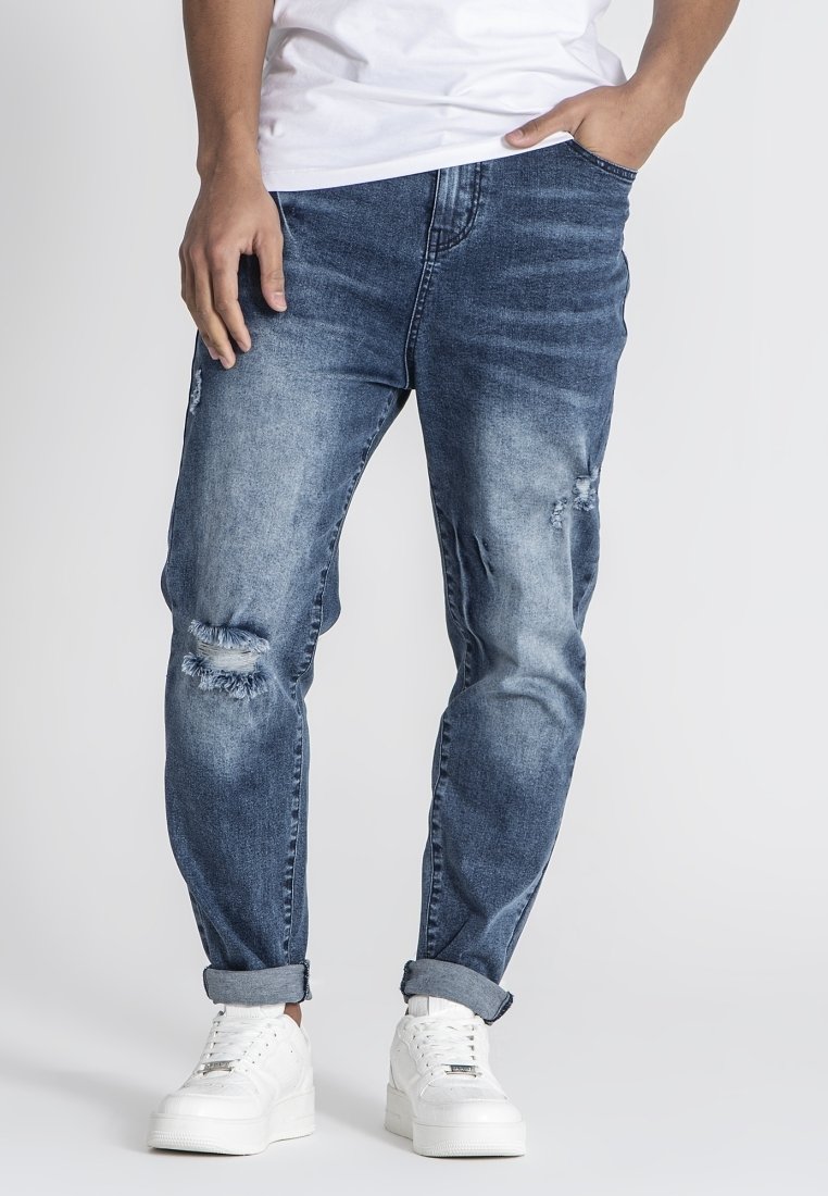 Jeans de mezclilla azul claro con desgastes, un corte relajado y puños enrollados, combinados con zapatillas blancas sobre un fondo liso.