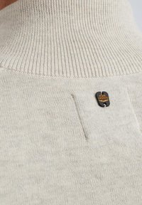 Helles beiges hochgeschlossenes Sweatshirt mit gerippter Textur, das auf der Rückseite in der Nähe einer Tasche ein kleines schwarzes Etikett mit orangefarbener Aufschrift trägt.