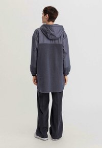 Giacca lunga grigia con cappuccio, con un retro simile alla lana e un frontale liscio. Presenta polsini elastici e orlo arrotondato. Abbinata a pantaloni scuri e sneaker.