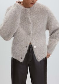 Person wearing a beige fuzzy buttoned cardigan and black leather pants, with one hand adjusting the collar.