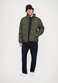 Casaco puffer verde-oliva com fecho na frente, apresentando um logótipo no peito, combinado com uma polo azul-marinho e calças azul-marinho. Ténis brancos completam o visual.