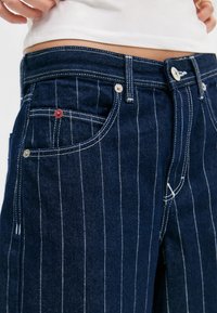 Jean en denim bleu avec des rayures verticales blanches, design à cinq poches, quincaillerie en argent et un accent logo rouge près de la poche avant.