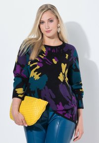 Frau mit langen blonden Haaren, die einen mehrfarbigen abstrakten Pullover und blaue Hosen trägt und eine strukturierte gelbe Clutch hält.