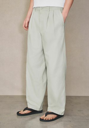 Person trägt lockere, hellgraue plissee Hose mit Seitentaschen und schwarzen Flip-Flops, steht auf einem grauen Boden vor einer beigen Wand.