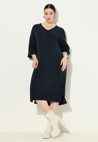 Abito lavorato a maglia blu navy con scollo a V, maniche tre quarti e spacchi laterali. Abbinato a stivali bianchi con tacco spesso. Texture liscia.