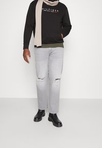 Felpa nera con logo, maglietta verde oliva sotto, jeans strappati grigio chiaro e stivaletti neri. Sciarpa che si avvolge attorno al collo.