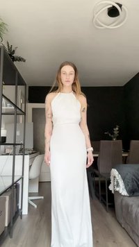 Fehér, nyakpántos ruha, amely testre simuló felsőrészt és uszályos szoknyát tartalmaz. Simává, bokáig érő anyag, egyszerű beltéri környezetben bemutatva.