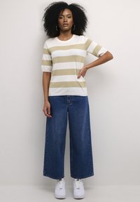 Gestreept gebreid topje in crème en beige met korte mouwen, gecombineerd met wijde blauwe spijkerbroeken en witte sneakers. De achtergrond is neutraal.