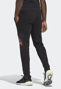 Černé tepláky vyrobené z měkkého materiálu, s bočními kapsami a velkou oranžovou nášivkou na dolní části nohy. Užší střih s elastickými manžetami.