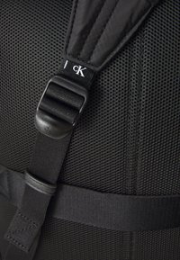 Black mesh backpack strap featuring plastic hardware and fabric accents, with visible stitching and brand label in a contrasting color.