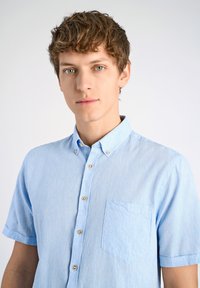 Ljusblå kortärmad skjorta med knäppning i texturerad tyg, med en bröstficka och kontrasterande knappar.