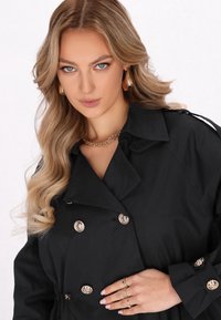 Černý trench coat s dvouřadým designem, zlatými knoflíky a velkým límcem. U stylu s vrstevnatým zlatým náhrdelníkem a prsteny.