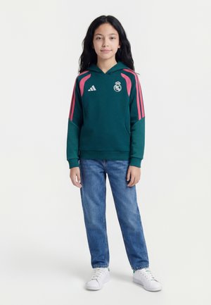 Enfant aux longs cheveux noirs portant un sweat à capuche Real Madrid vert foncé et rose, jean bleu et baskets blanches, debout face à l'avant.
