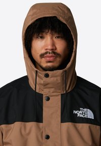 Brązowa i czarna kurtka z kapturem, z przednim zapięciem na napy i logo. Gładka tekstura z designem w kolorowe bloki.