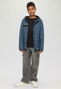 Blauwe geïsoleerde jas met een capuchon, gewatteerde voorkant, zwarte accenten, gedragen over een zwart shirt en baggy grijze cargo-broek, gecombineerd met witte sneakers.