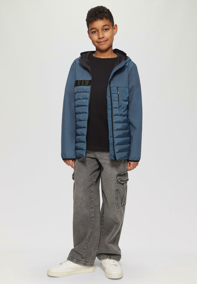 Blauwe geïsoleerde jas met een capuchon, gewatteerde voorkant, zwarte accenten, gedragen over een zwart shirt en baggy grijze cargo-broek, gecombineerd met witte sneakers.