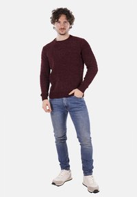 Uomo con capelli ricci che indossa un maglione a maglia di colore bordeaux, jeans blu e sneakers bianche, in piedi con una mano in tasca.