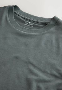 Nahaufnahme eines grauen, körperbetonten T-Shirt-Ausschnitts, zeigt das Größenschild Medium und den Schriftzug "Made in Bangladesh" im Kragen.