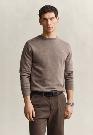 Junger Mann mit kurzen lockigen Haaren, der einen enganliegenden braunen Pullover, passende Hosen, einen schwarzen Gürtel und eine Armbanduhr trägt, vor einem einfarbigen Hintergrund steht.