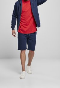 Haut zippé bleu marine avec poches, associé à un T-shirt rouge et un short bleu marine. Des baskets blanches complètent le look sportif.