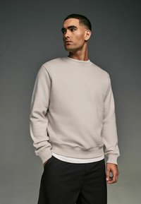 Sudadera beige clara con cuello redondo, puños acanalados y dobladillo, combinada con pantalones cortos negros, sobre un fondo oscuro.